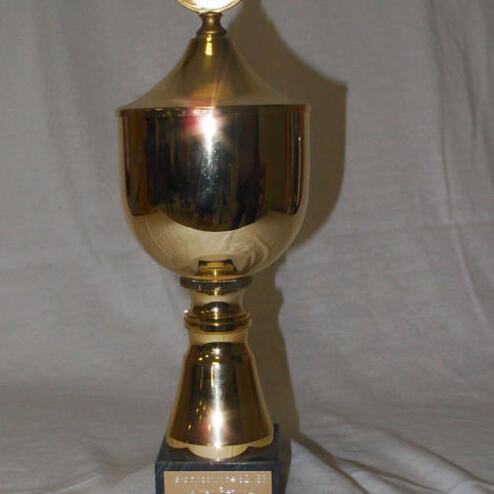 Goldener Pokal mit rundem Deckel und schwarzem Sockel, gravierte Plakette auf der Vorderseite.