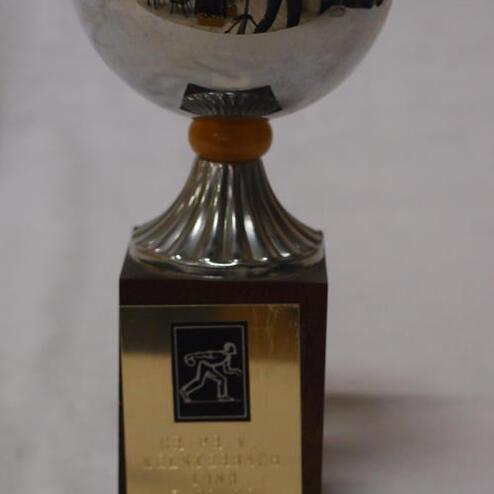 Silberner Pokal mit goldener Schale auf einem braunen Sockel, graviert mit einem Sportmotiv und einem Jahresdatum.