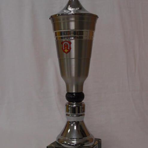 Silberner Pokal mit Wappen und Sportmotiv auf einem schwarzen Sockel. Beschriftung auf der Basis sichtbar.
