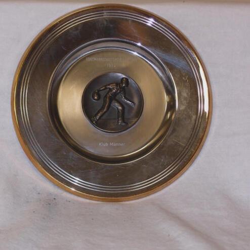 Silberne Schüssel mit einer Bronze-Plakette eines Sportlers, der einen Ball wirft, auf einem hellen Untergrund.