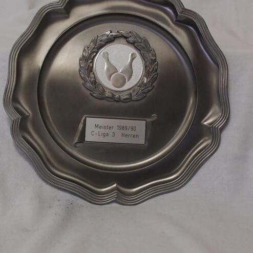 Silberner Meisterschaftsteller mit Wappen und der Inschrift „Meister 1989/90 C-Liga 3 Herren“ auf weißem Hintergrund.