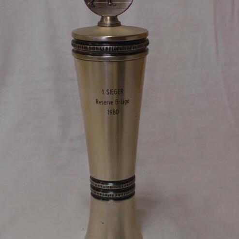 Siegertrophäe aus Metall mit Inschrift „1. SIEGER Reserve B-Liga 1980“ und figurativem Pokalaufsatz.
