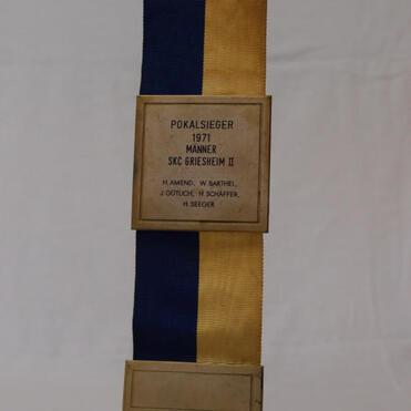 Pokal in gold, blau-gelber Stoffband mit Inschrift "Pokalsieger 1971 Männer SKC Griesheim II" und Namen.