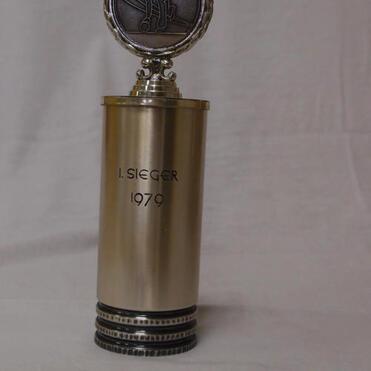 Pokaltrophäe mit der Aufschrift „I. SIEGER 1979“ und einer Medaille an der Spitze, auf einem weißen Hintergrund.
