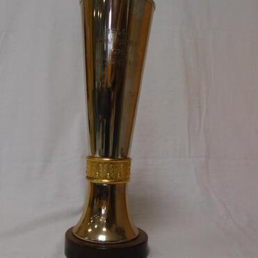 Goldener Pokal mit flachem Deckel, graviert, auf einem runden Holzsockel, vor einem weißen Hintergrund.