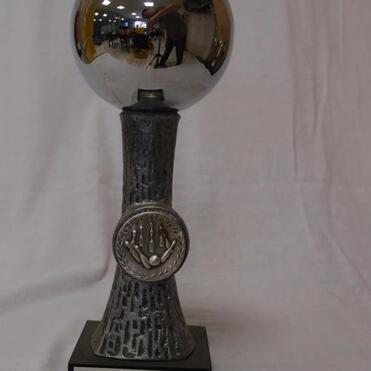 Pokalschale mit reflektierender Oberfläche und gravierter Siegerplakette, auf grauem Sockel mit Beschriftung.