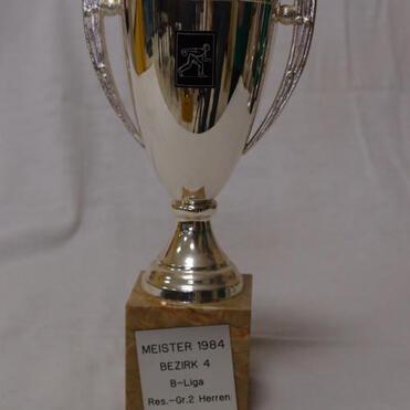 Silberner Pokal mit Griffen und einer Inschrift: „Meister 1984 Bezirk 4 B-Liga Res.-Gr. 2 Herren“ auf einem Sockel.