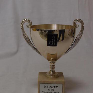 Goldener Pokal mit zwei Henkel, beschriftet mit "MEISTER 1986 Vereinsklasse SKC IV" auf einem Holzsockel.