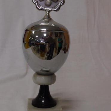 Schimmernder Sportpokal auf schwarzem Sockel mit gravierter Inschrift und Zinnmedaille mit Sportmotiven oben.