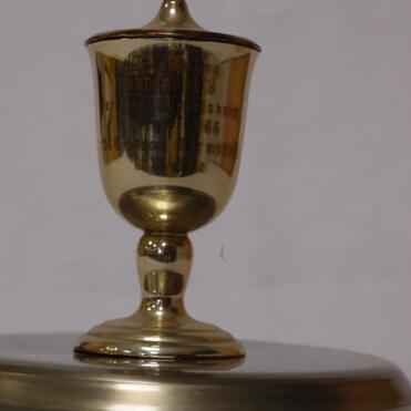 Goldener Pokal auf einem silbernen Untersetzer, mit eingravierter Beschriftung, leicht schräg abgebildet.