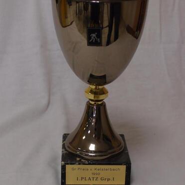 Silberne Pokalschale auf schwarzem Sockel mit der Aufschrift „1. PLATZ Grp.1, Gr. Preis v. Kelsterbach 1990“.