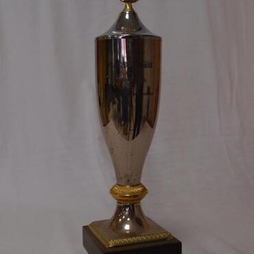 Silberner Pokal mit goldener Verzierungen auf einem dunklen Sockel, präsentiert auf einem neutralen Hintergrund.