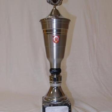 Silberner Pokal mit einem Symbol und einer Gravur auf schwarzem Sockel, auf hellem Hintergrund.