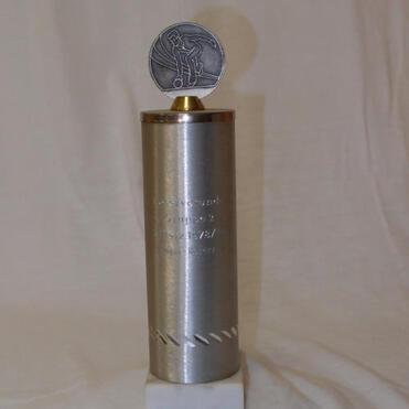Silberner Pokal mit kreisförmiger Medaille oben, auf weißem Sockel, mit eingraviertem Text.
