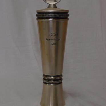 Pokale mit Aufschrift „1. Sieger Reserve A-Liga 1980“ und einer Sportlerfigur als Deckel auf grauem Hintergrund.