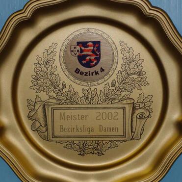 Goldene Ehrenplakette mit Aufschrift "Meister 2002 Bezirk 4 Damen" und dekorativen Elementen, an einer blauen Wand aufgehängt.