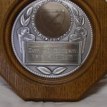 Plakette aus Holz mit silberner Medaille, verzierte Ränder und Inschrift „Zum Jubiläum Vereinsgründung“.