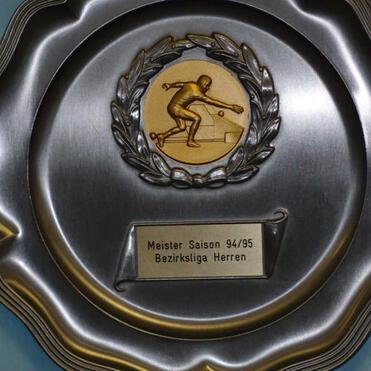 Silbernes Meisterschaftsschild mit einer goldenen Medaille und der Aufschrift „Meister Saison 94/95 Bezirksliga Herren“.