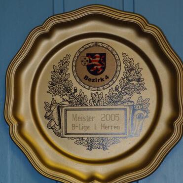 Goldene Meisterschaftsplakette von 2005 für B-Liga Herren, verziert mit einem Wappen und Pflanzenmotiven.