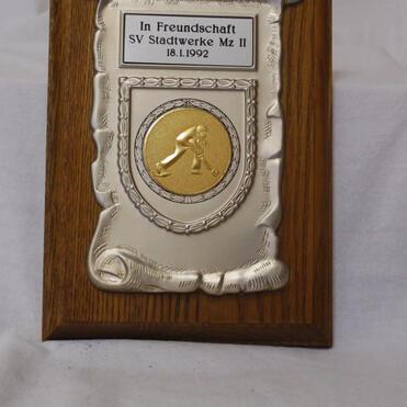Ehrenplaque aus Holz mit silbernem Schild und goldener Medaille, beschriftet mit „In Freundschaft SV Stadtwerke Mz II 18.1.1992“.