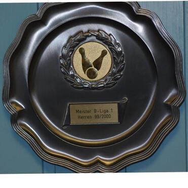 Plakette mit goldener Auszeichnung für Meisterschaft in B-Liga 1 Herren 99/2000, im Hintergrund eine blaue Wand.