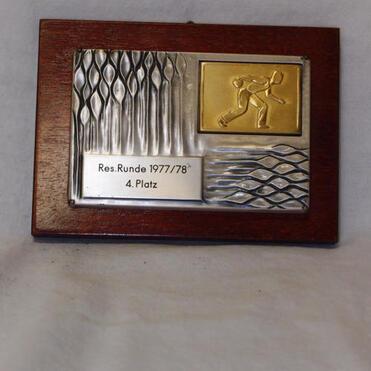 Holzplatte mit einer Metallauszeichnung, graviert mit "Res. Runde 1977/78" und "4. Platz", zeigt einen sportlichen Bewegungsablauf.