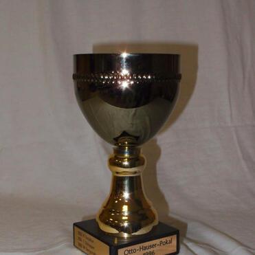 Silberner Pokal auf schwarzem Sockel mit der Aufschrift „Otto-Hauser-Pokal 1996“. Hintergrund ist ein graues Tuch.