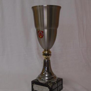 Silberne Trophäe mit goldenem Sockel und Wappen auf schwarzem Grund, steht auf einem hellen Untergrund.