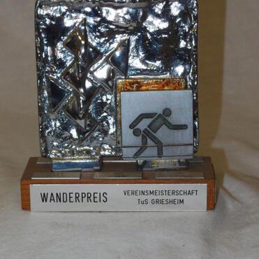 Wanderpreis aus Metall mit stilisierten Sportmotiven und Gravur „Vereinsmeisterschaft TuS Griesheim“ auf Holzsockel.