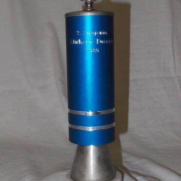 Silberner Pokal mit blauer Figur, gravierten Texten und runder Fußplatte aus Marmor.