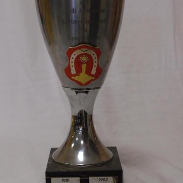 Silberner Pokal mit Wappen und Gravur, beschriftet mit „1981 TUS Olympia“ und „1982 TUS Olympia“ auf schwarzem Sockel.