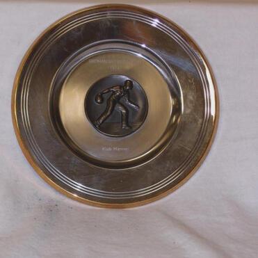 Silberne Schüssel mit einer Bronze-Plakette eines Sportlers, der einen Ball wirft, auf einem hellen Untergrund.