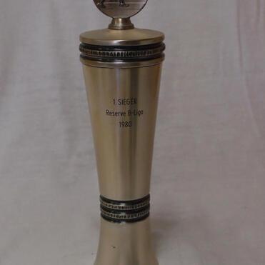 Siegertrophäe aus Metall mit Inschrift „1. SIEGER Reserve B-Liga 1980“ und figurativem Pokalaufsatz.