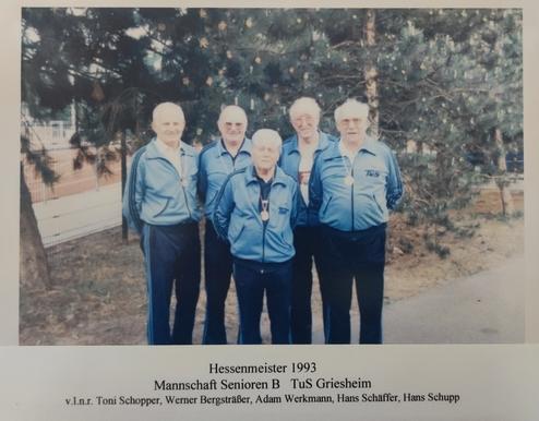 Fünf Senioren in blauen Trainingsanzügen stehen lächelnd vor einer grünen Wand mit Bäumen, sie tragen Medaillen.