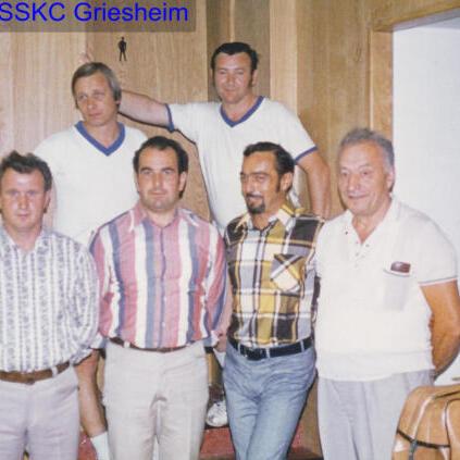 Gruppe von sieben Männern unterschiedlichen Alters in sportlicher Kleidung vor einer Holzvertäfelung.