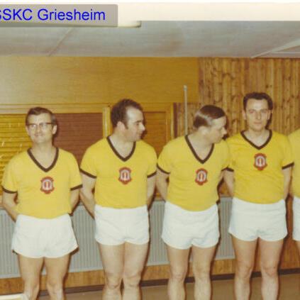 Fünf Männer in gelben Shirts und weißen Shorts stehen aufgereiht vor einer Holzvertäfelung.