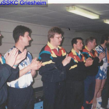 Acht Männer in Sportjacken applaudieren in einer Sporthalle, im Hintergrund eine neutrale Wand.