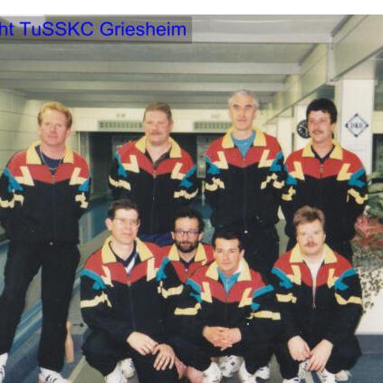 Foto einer Gruppe von acht männlichen Sportlern in bunten Trainingsanzügen, auf einer Kegelbahn posierend.