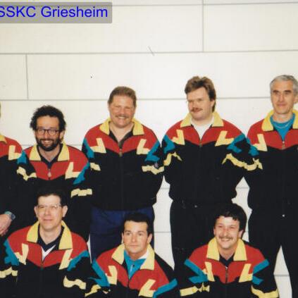 Gruppenfoto von acht Männern in bunten Sportjacken, lächelnd vor einer hellen Wand.