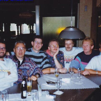 Gruppe von sechs Männern an einem Tisch in einem Restaurant, umgeben von Gläsern und einer Lampe im Hintergrund.