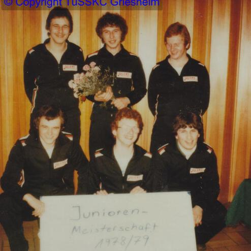 Sechs männliche Sportler in Trainingsanzügen posieren vor Holzpaneelen mit einem Schild zur Junioren-Meisterschaft 1978/79.