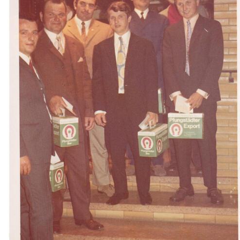 Gruppe von Männern in formeller Kleidung, die Kisten mit der Aufschrift „Plungstädter Export“ halten.