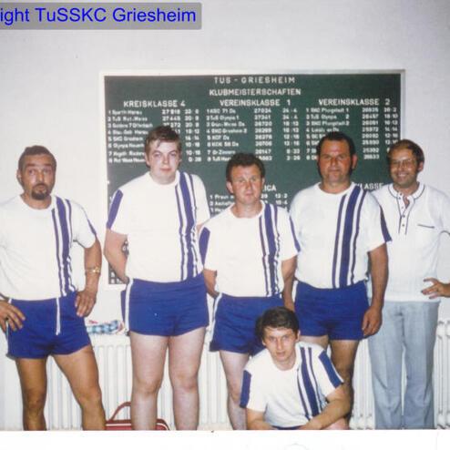 Gruppenfoto von sechs Sportlern in einheitlicher Kleidung vor einer Ergebniswand in einem Vereinsraum.