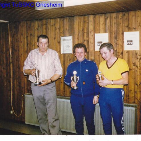 Drei Männer halten Pokale in einem Holzraum; an der Wand hängen Urkunden.