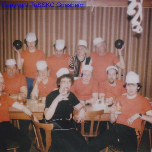 Gruppe von zehn Personen in roten T-Shirts und weißen Hüten, stehend und sitzend um einen Tisch mit Getränken.