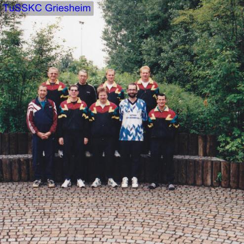 Gruppenfoto von acht Männern in Sportanzügen, die auf einem gepflasterten Platz mit Bäumen stehen.