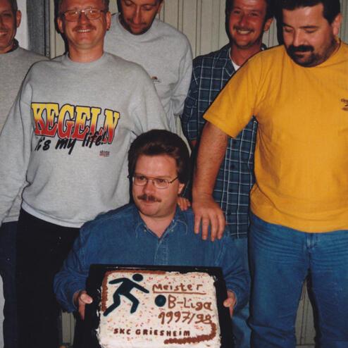Sechs Männer stehen und sitzen um einen Mann, der eine Torte mit der Aufschrift „Meister B-Liga 1997/98“ hält.