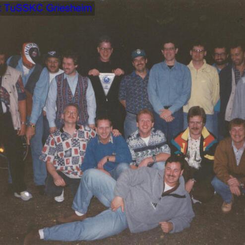 Gruppenselfie von Männern in sportlicher Kleidung, lächelnd vor einem dunklen Hintergrund, einige sitzen und stehen.