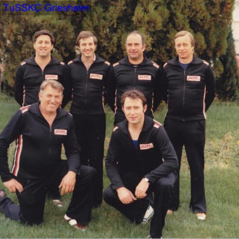 Sechs Männer in Sportanzügen posieren lächelnd im Freien vor einem grünen Hintergrund mit Sträuchern und Bäumen.