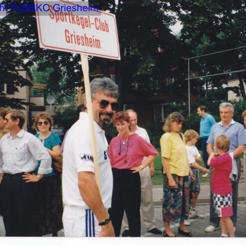 Mann mit Sonnenbrille hält ein Schild mit der Aufschrift „Sportkegel-Club Griesheim“ in einer Gruppe von Menschen.